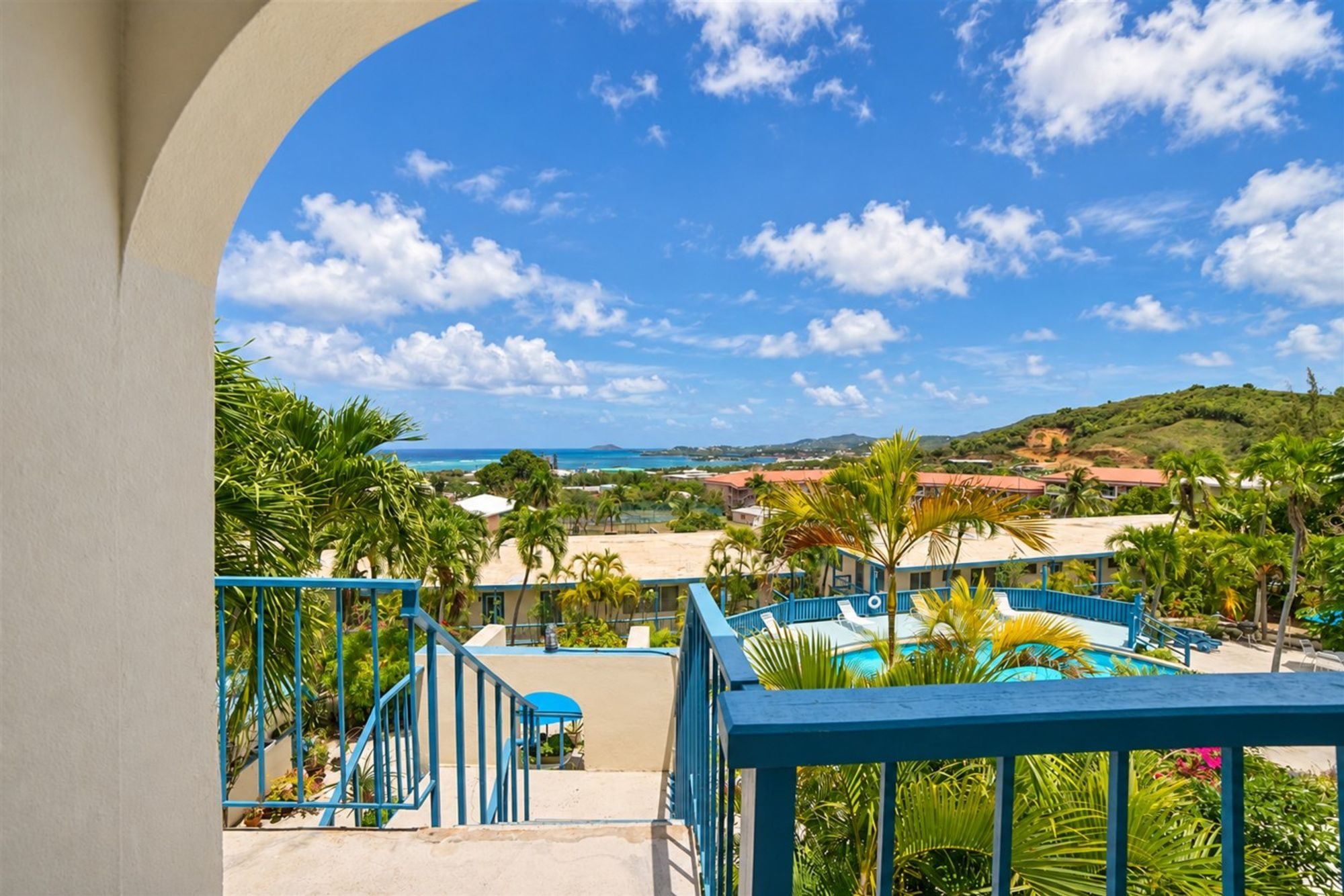 View through a stucco arch at the top of blue-railed stairs, over palms and a pool to green hills and the sea