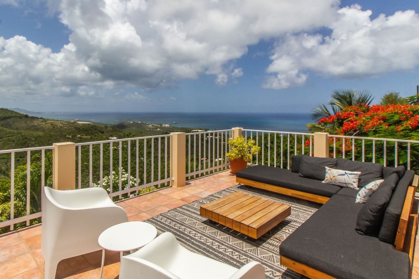 Terrace with a dark outdoor sectional, white chairs and a teak table, red flowering trees and the sea beyond the railing