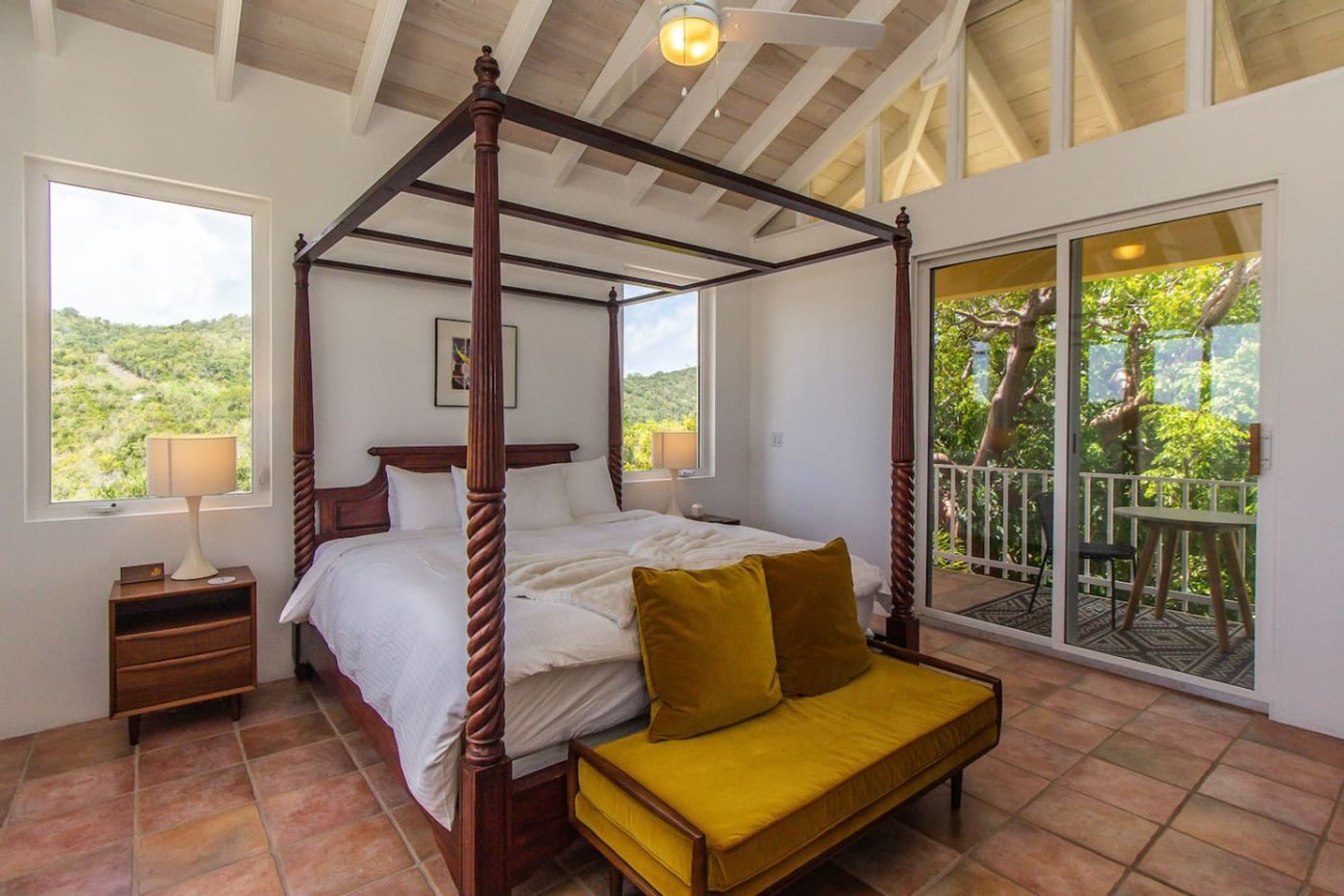 Bedroom with a dark four-poster bed, a mustard bench and glass doors onto a balcony over green hills