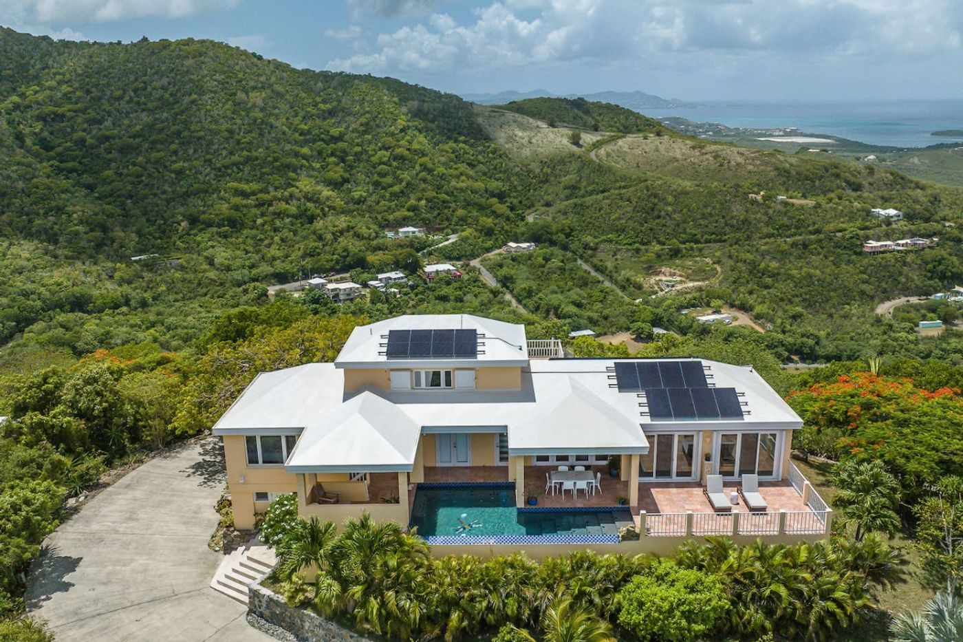 Aerial view of a pale villa with solar panels and a pool on a forested hilltop, the sea in the distance