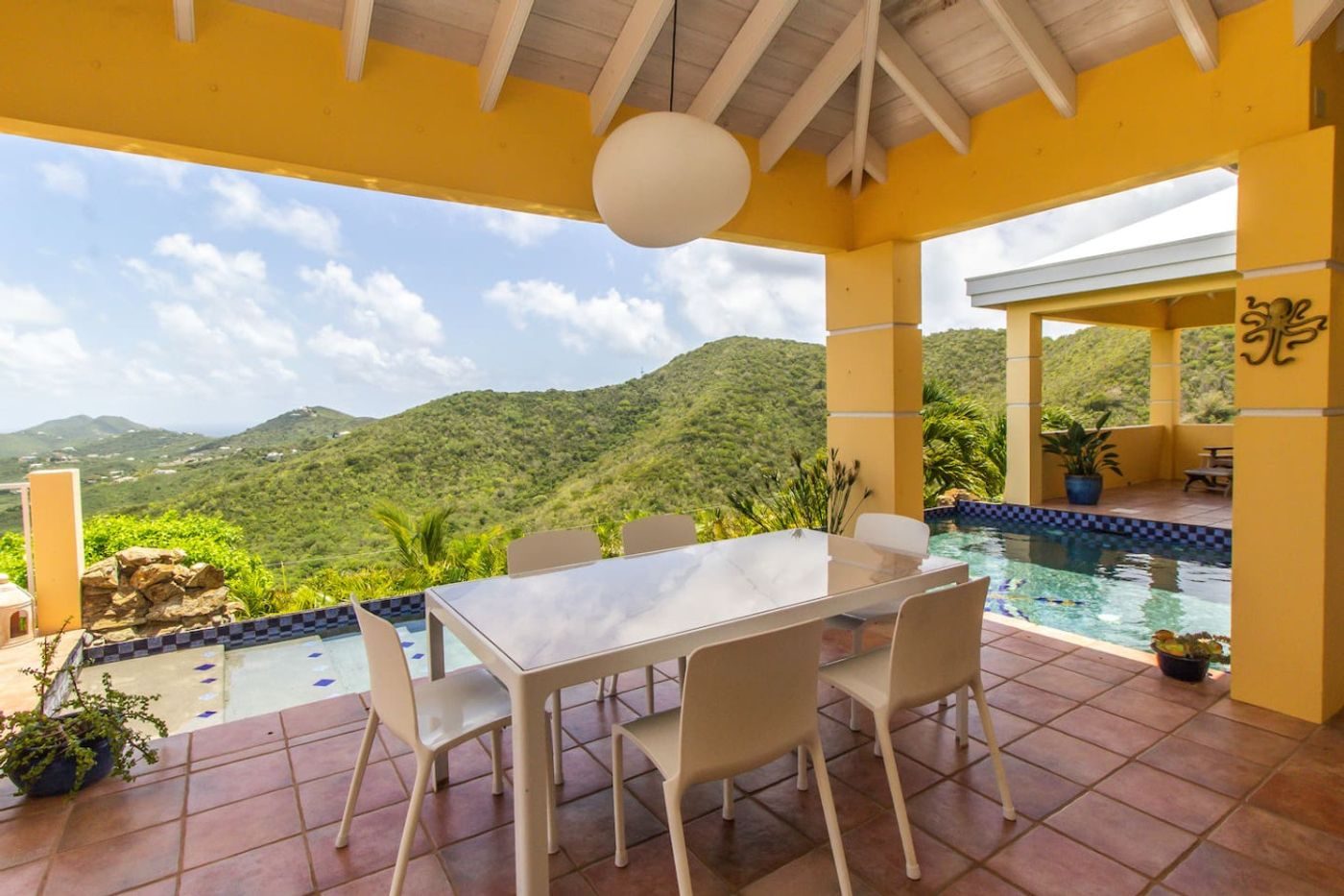 Covered terrace with a white dining table and chairs beside the pool, green hills beyond