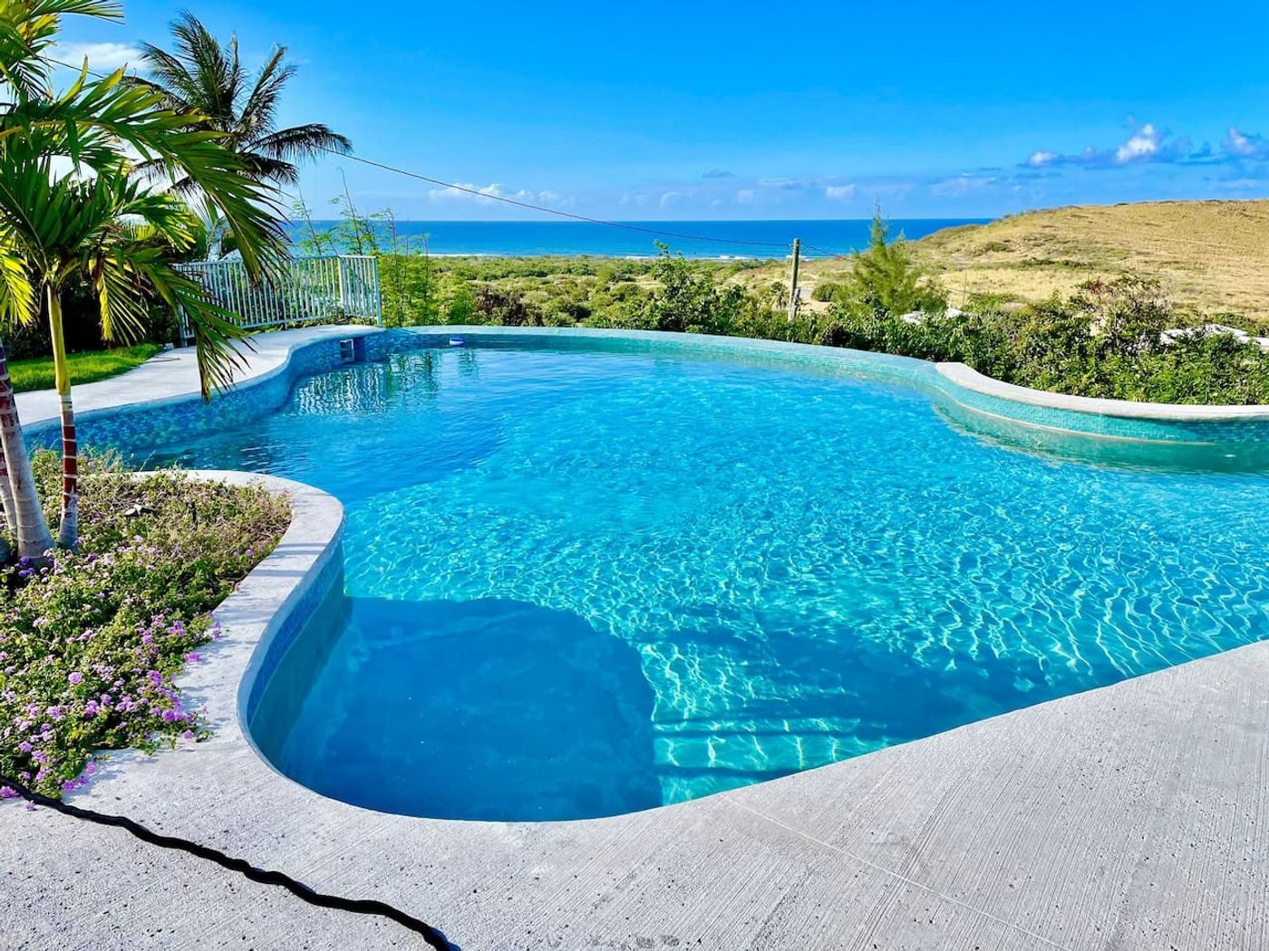 Curving turquoise pool edged with palms and flowering plants, with dry hills and the sea on the horizon