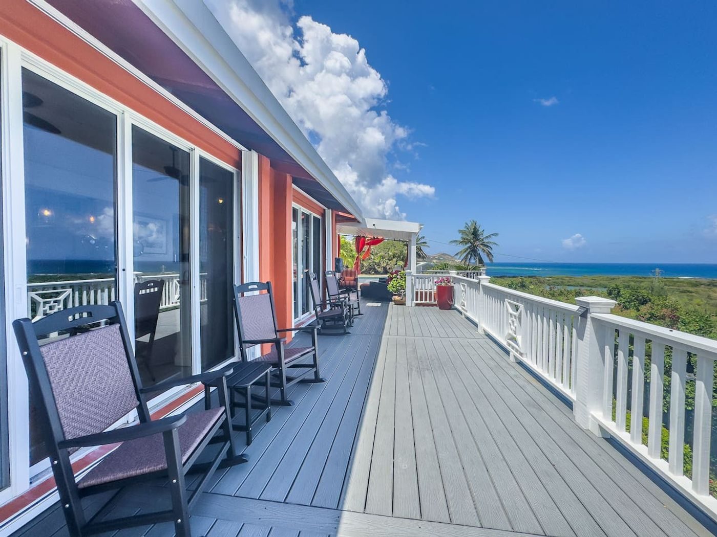 Long balcony with white railings and rocking chairs beside the orange-painted house, looking out to sea