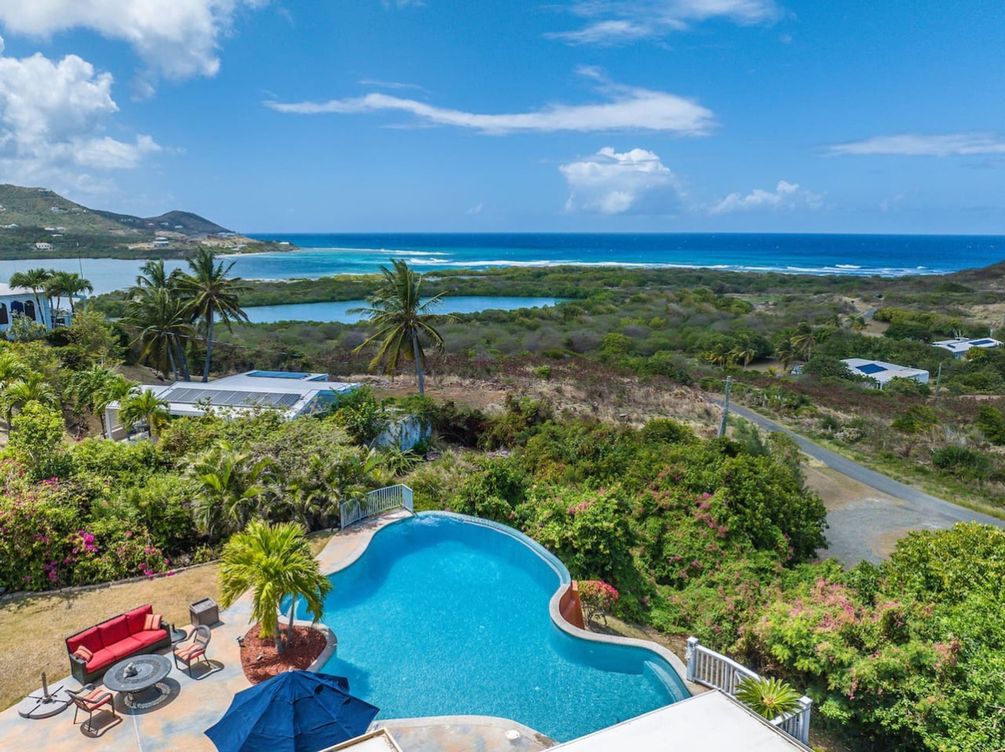 Aerial view of a curving pool and patio among tropical planting, with a bay, green hills and the open sea beyond