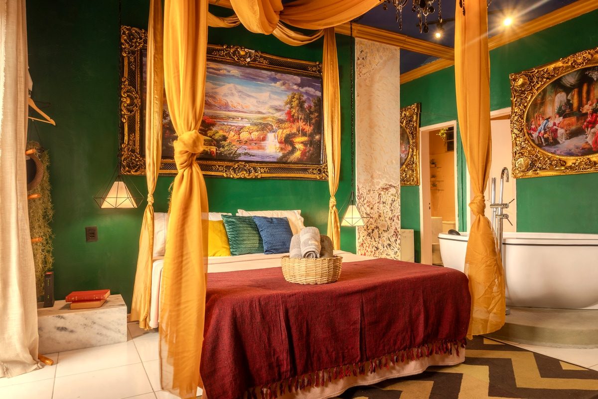 Canopy bed hung with gold curtains under a large gilt-framed landscape painting, a red throw and blue and yellow cushions