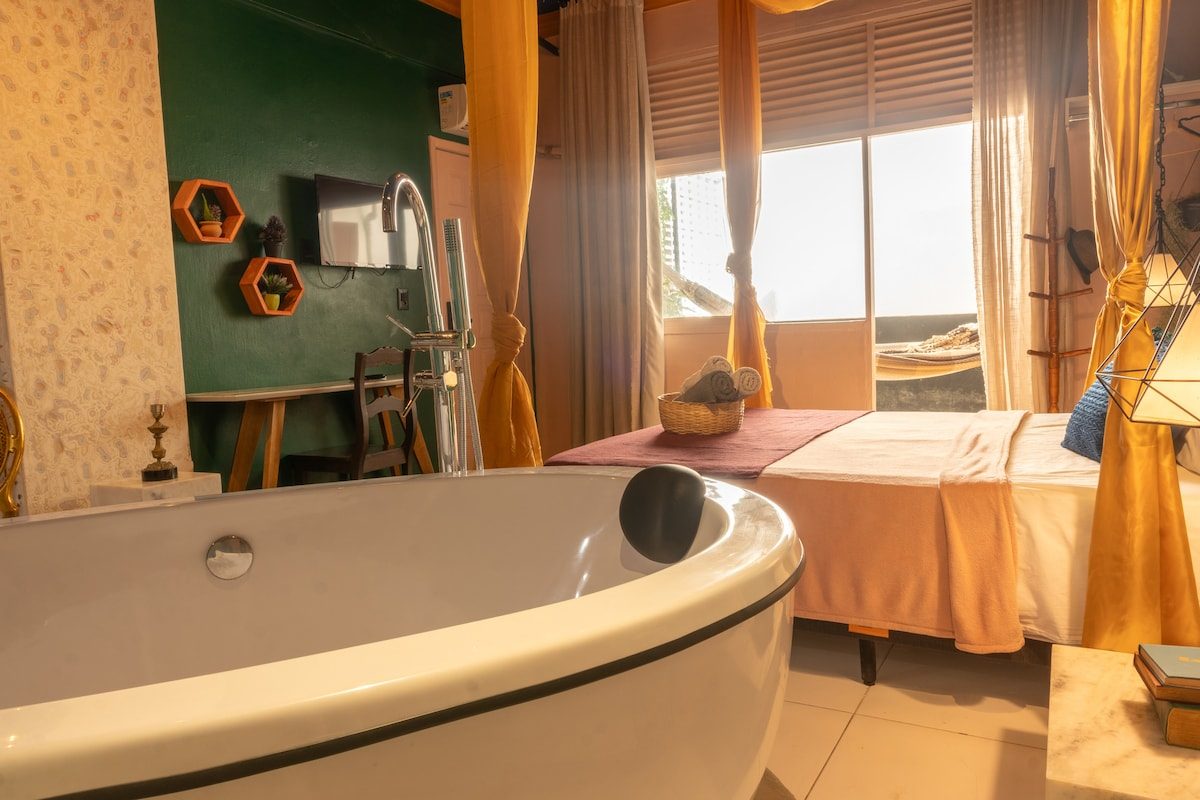 Freestanding tub by a window with gold curtains, the bed and a desk behind it