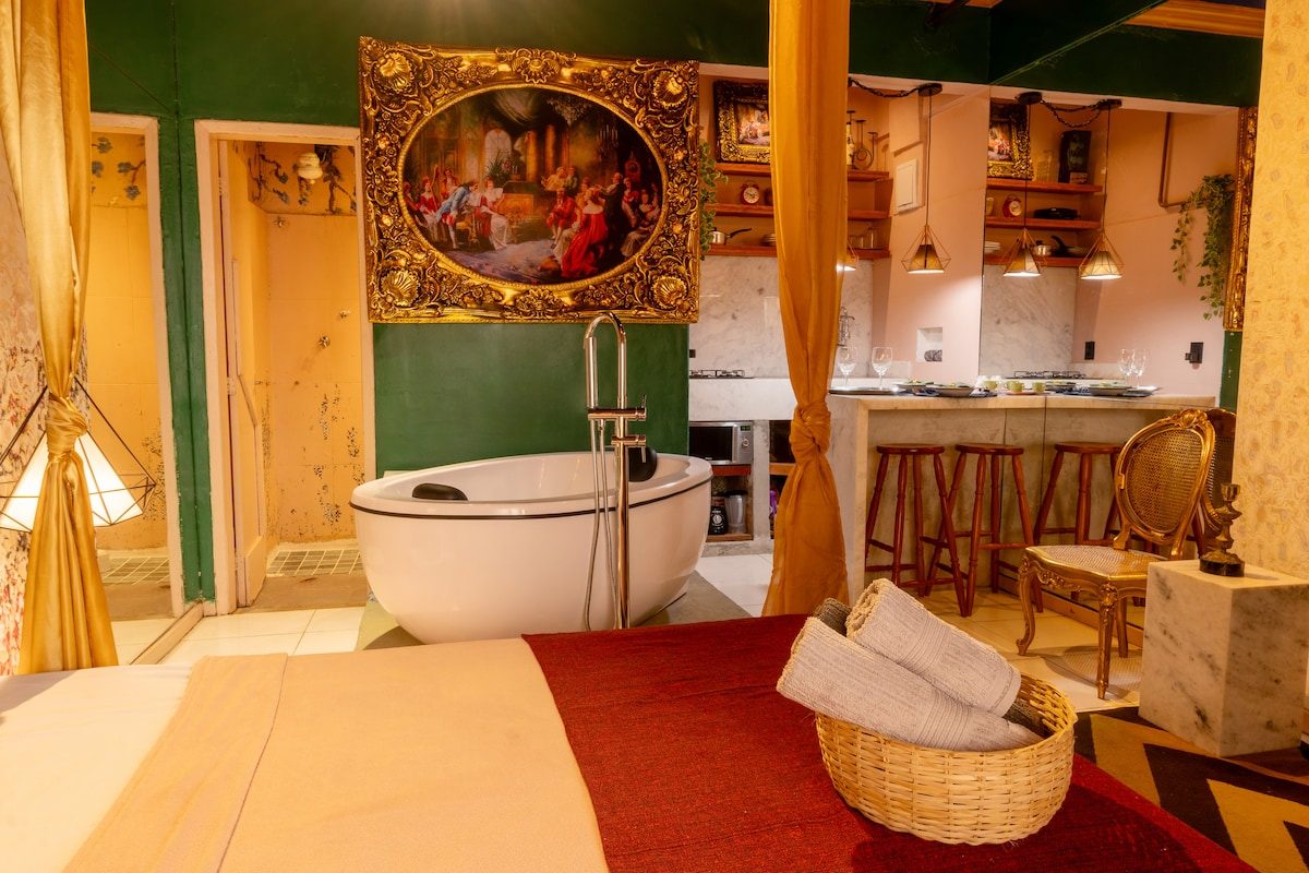 Loft with emerald walls and gold curtains, a freestanding white tub, a gilt-framed painting and a kitchen bar with stools beyond