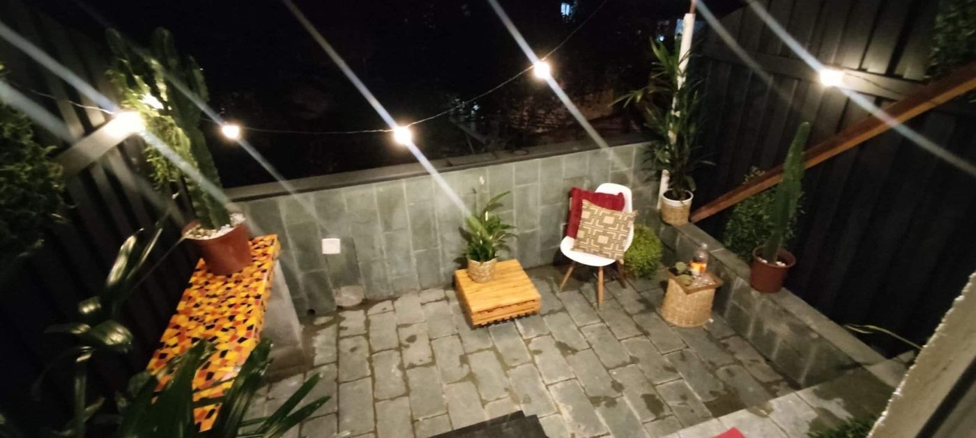 The terrace at Lux Vidigal at night, lit by a string of bulbs over a stone floor, with a white chair with red and patterned cushions, a low wooden table, a mosaic-tiled bench and tall potted plants