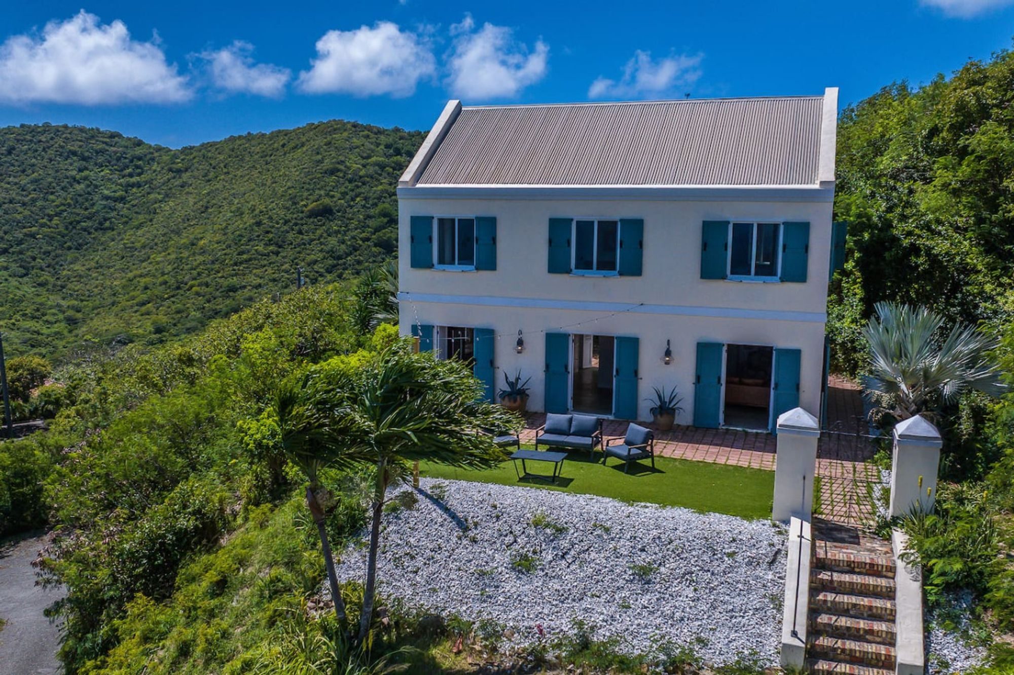 A two-storey white West Indian-style house with blue shutters and a grey roof on a green hilltop