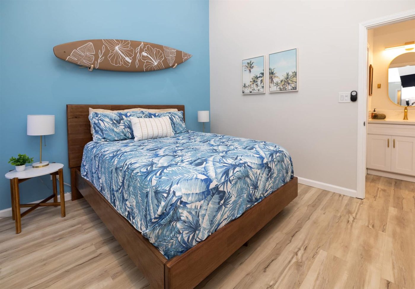 A bedroom at Sea Sun Maui with a queen bed in a dark wood frame under a blue leaf-print cover, a white lamp on a table at each side, a surfboard on the blue wall and a door through to the bathroom