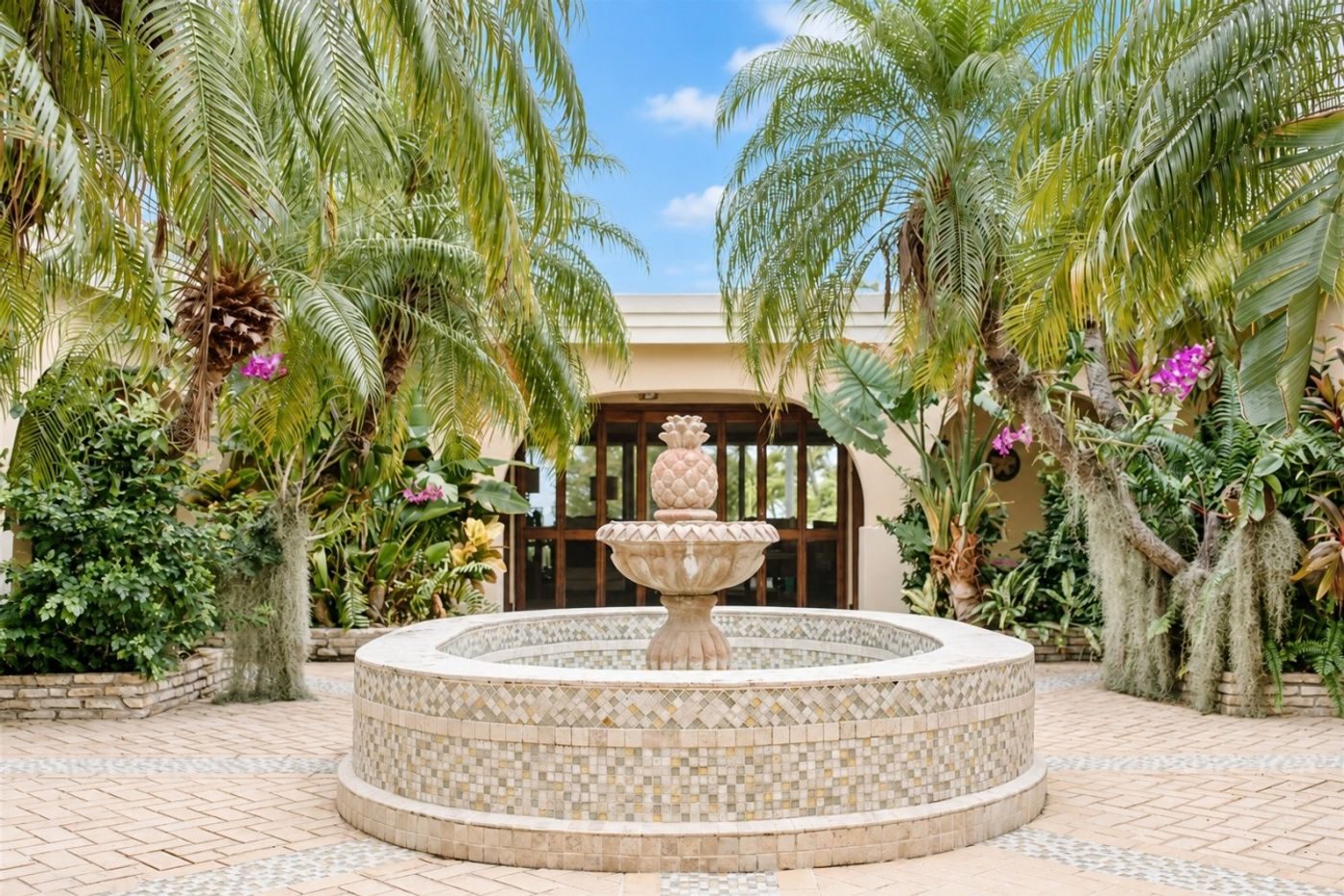 Stone fountain in a paved courtyard surrounded by palms and flowering plants