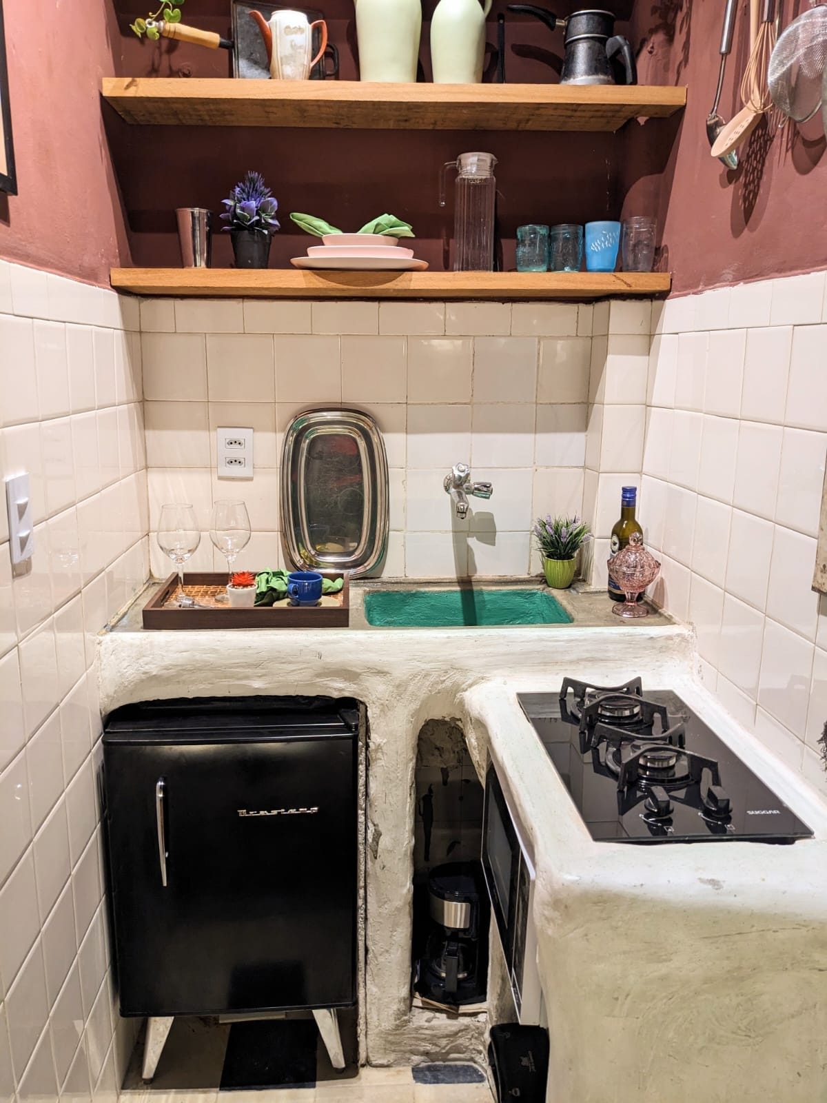 Small white-tiled kitchen with a two-burner stove, a mini fridge, wooden shelves of jars and a sink