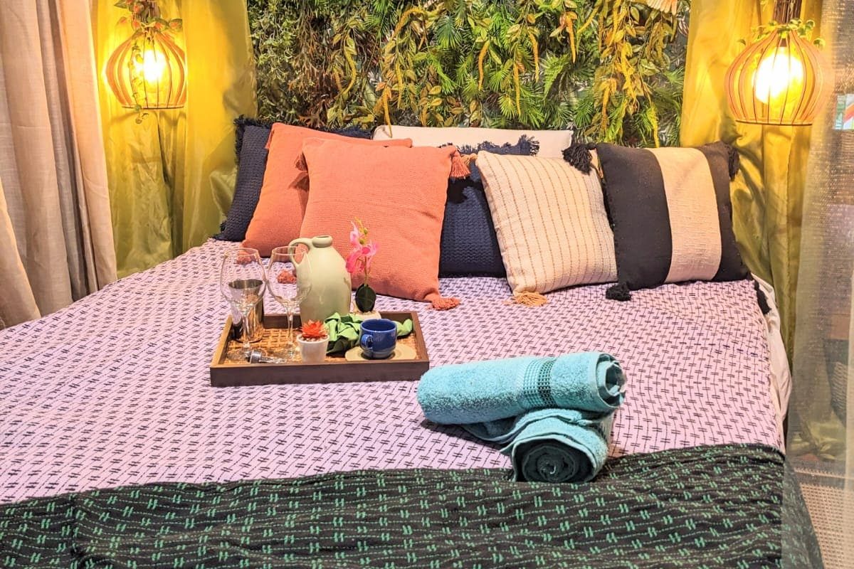 Bed with a lilac patterned cover, coral and navy cushions and a tray, under a wall of green hanging plants and warm lamps