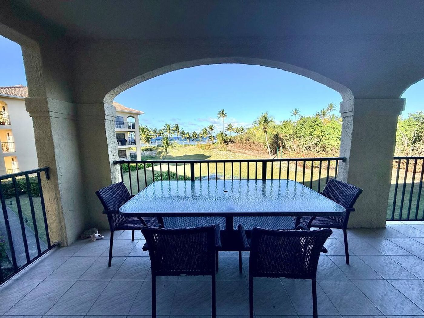 Covered balcony with a glass table and four chairs, an arched opening onto palms and blue sky
