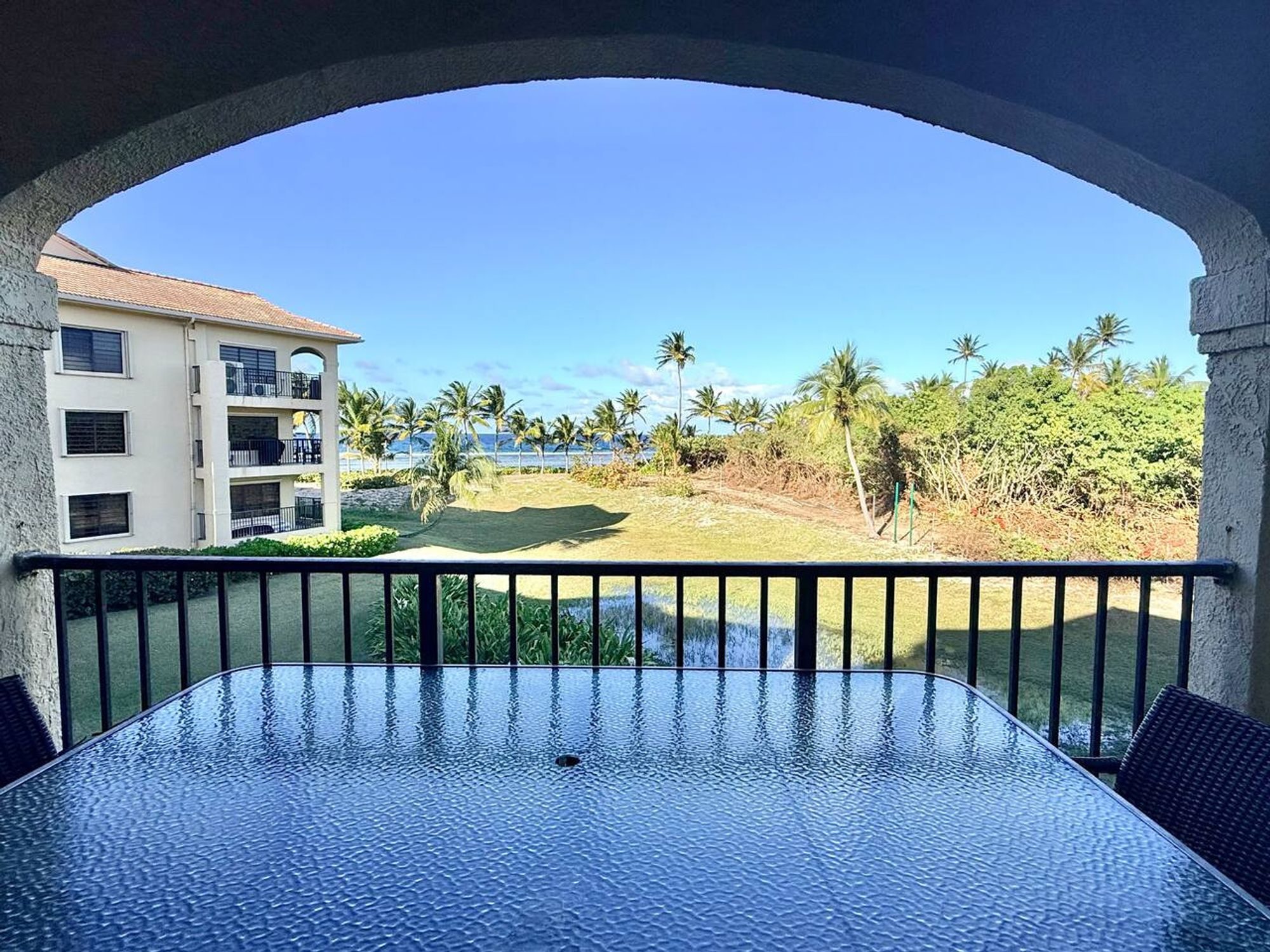 View through a stucco arch from a covered balcony over a glass dining table to lawns, palms and a white condominium building