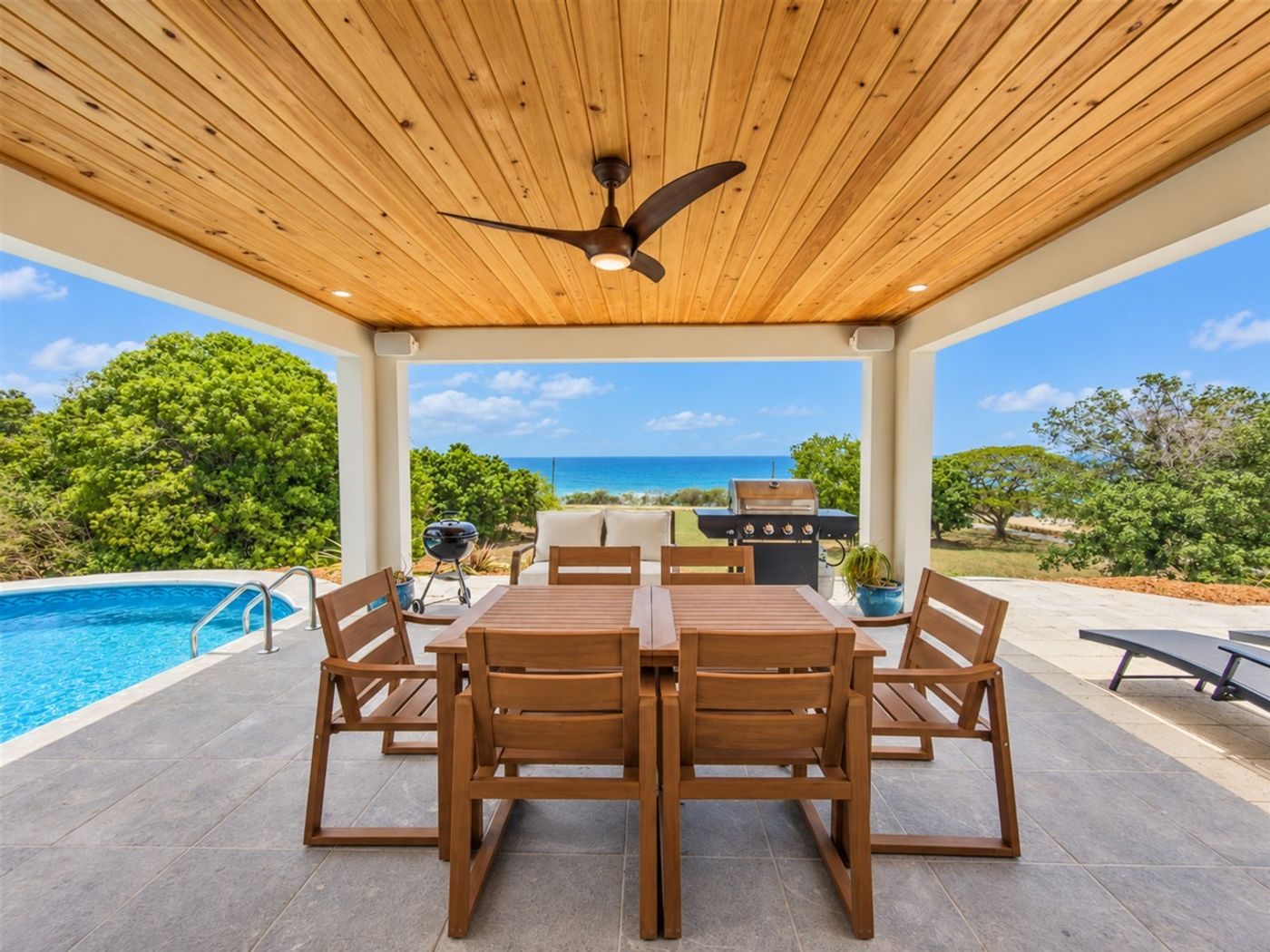 Covered terrace with a wood-plank ceiling and a teak dining table, the pool and the sea beyond