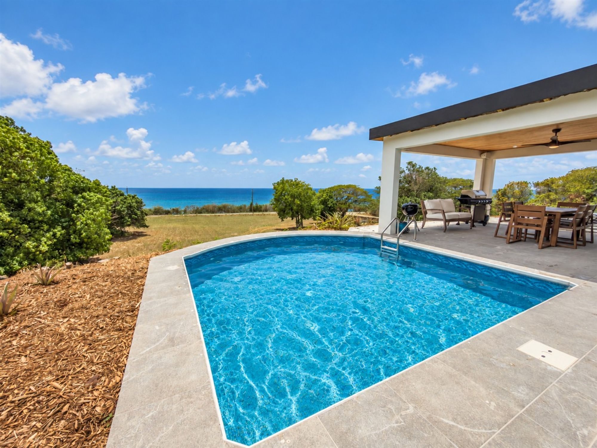 Private pool on a stone deck beside a covered terrace, with scrub trees and the sea on the horizon