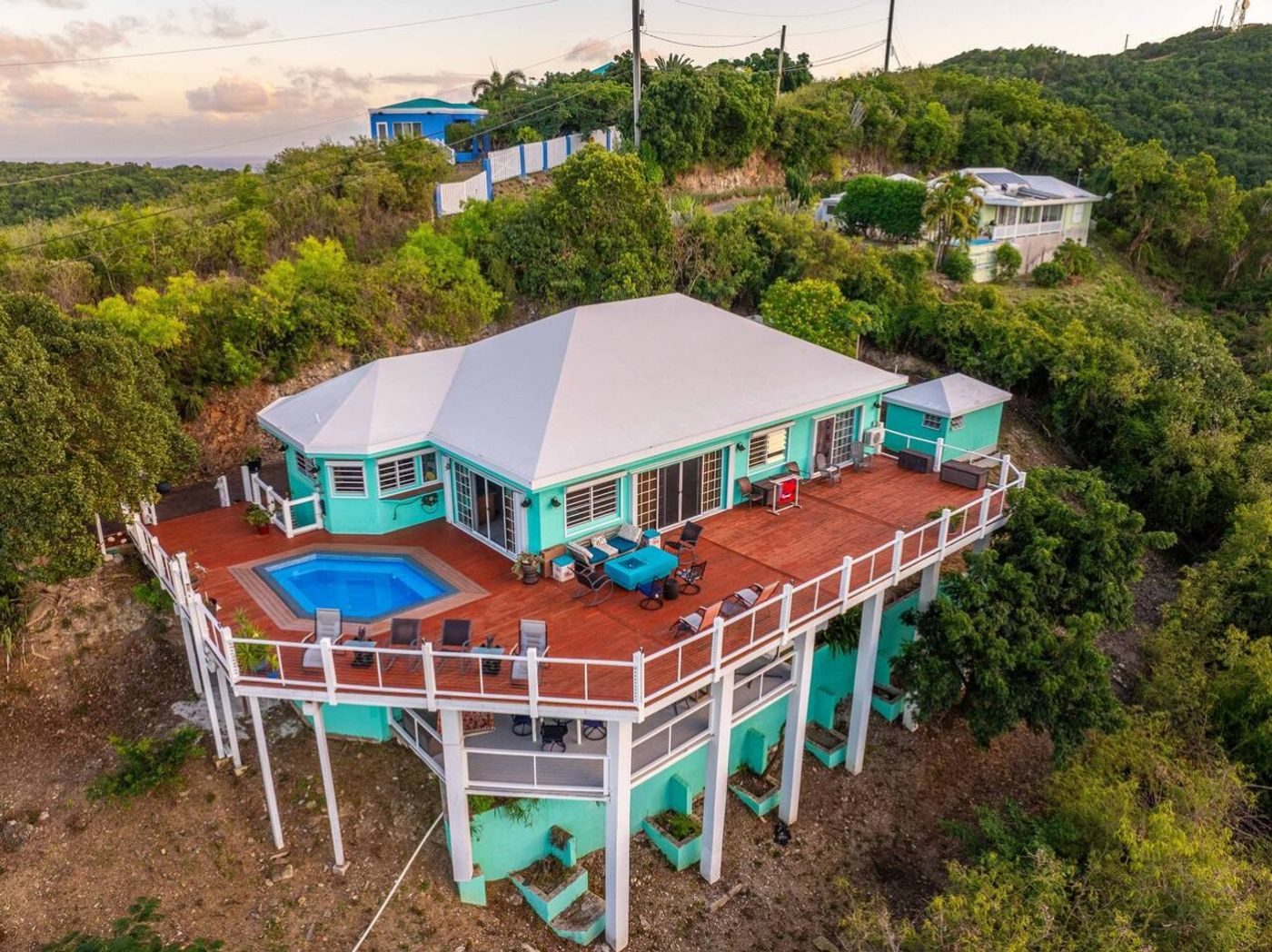 Aerial view of a turquoise-painted house with a white roof and a wraparound deck and pool on a wooded hillside