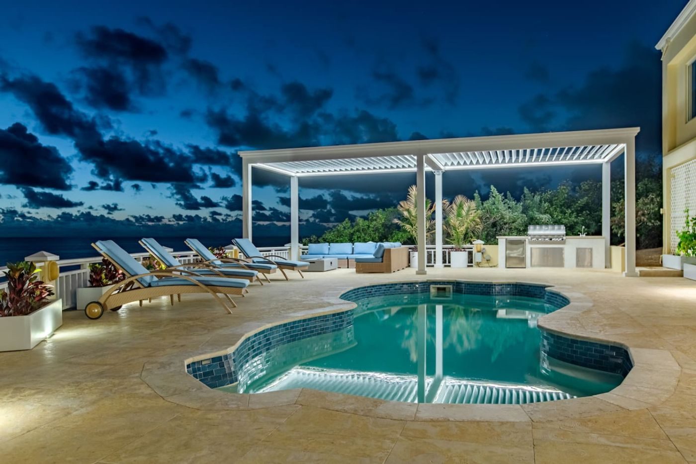 The free-form pool lit at twilight, with loungers and a white pergola over the outdoor kitchen