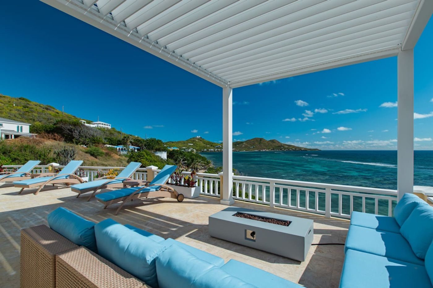 Covered terrace with a blue sectional and a fire table under a louvred pergola, looking over the sea to green hills