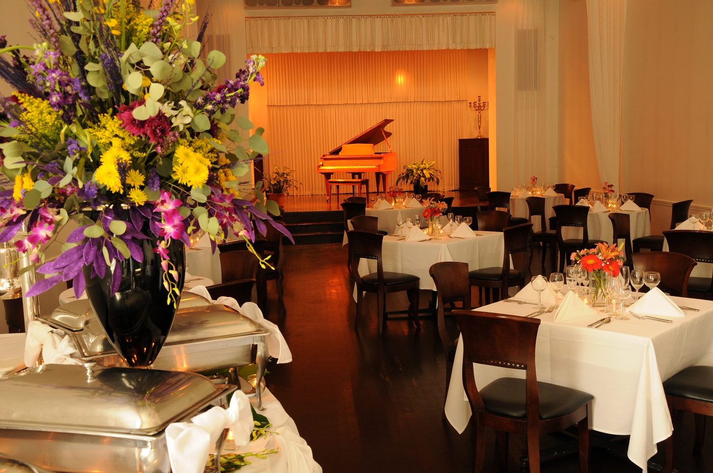 Round tables dressed in white linen before a lit grand piano stage in the Old Eagle Room at The Polo Inn
