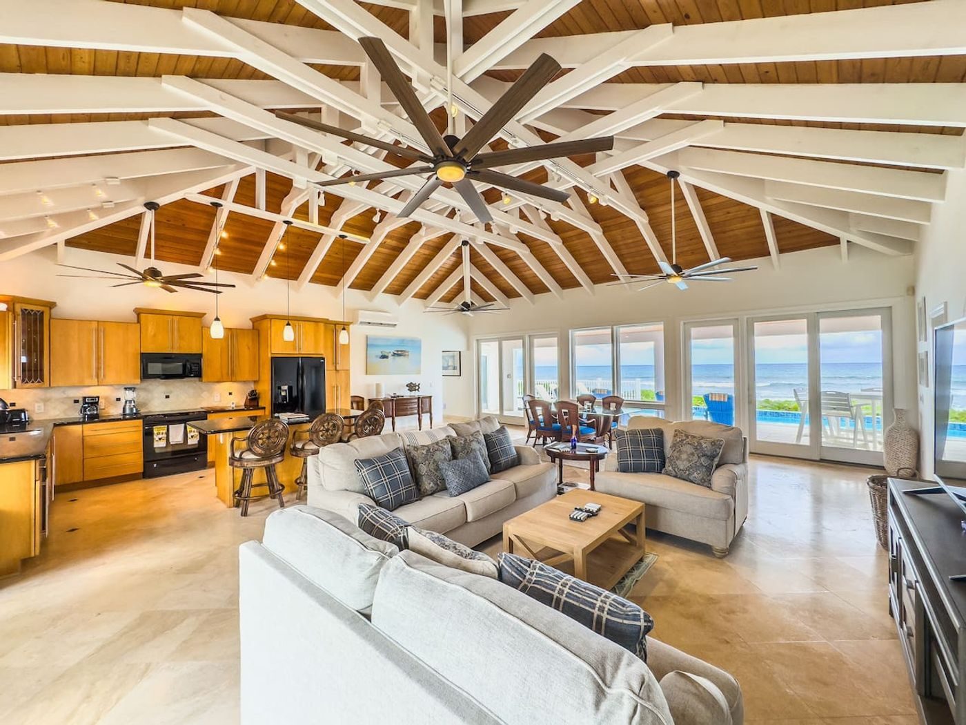Open living, dining and kitchen area under a high pitched ceiling of wood boards and white beams, with windows to the sea