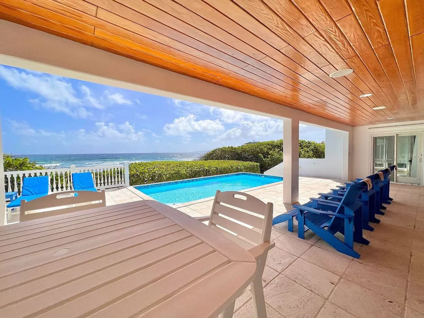 Covered gallery with an outdoor dining table and blue Adirondack chairs looking out over the pool to the sea