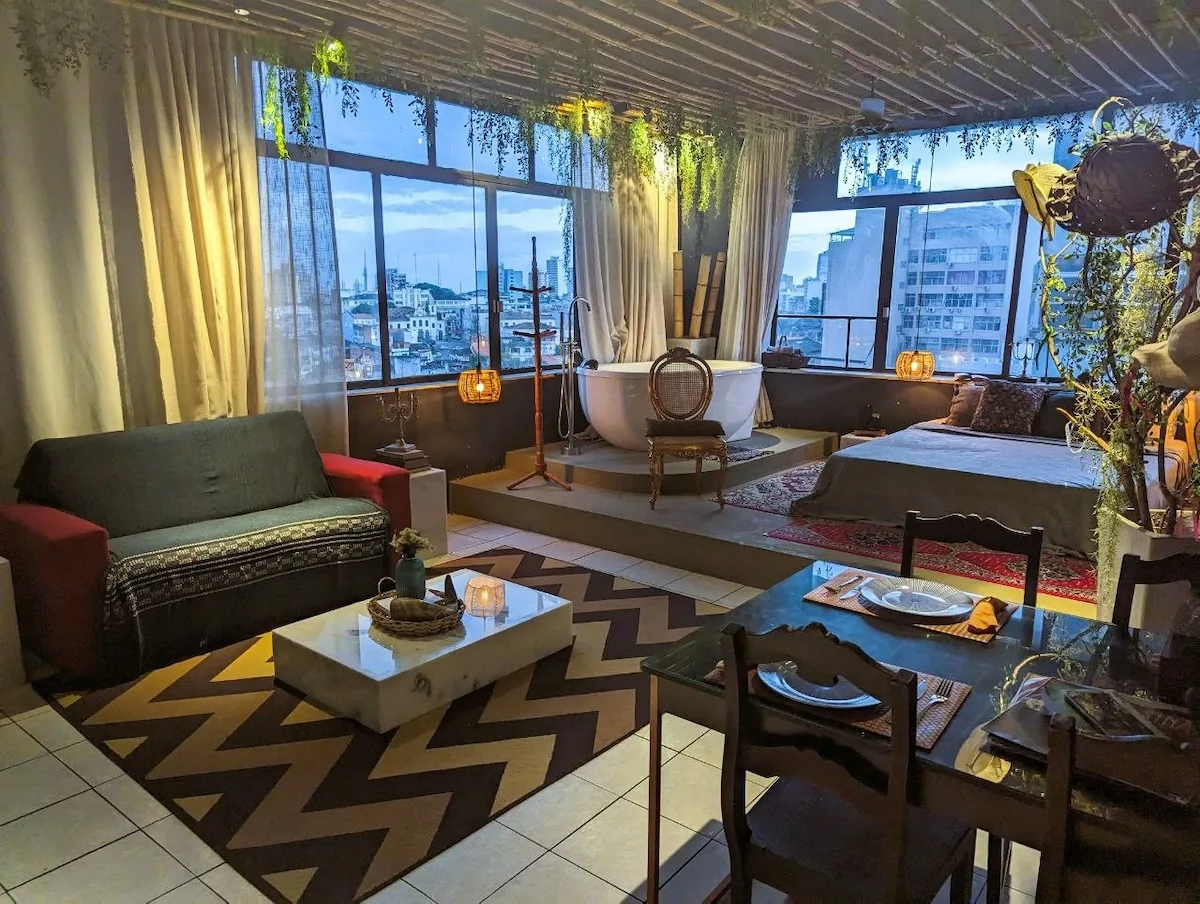 The Penthouse Jacuzzi loft at dusk — a freestanding tub on a platform by floor-to-ceiling windows over Salvador
