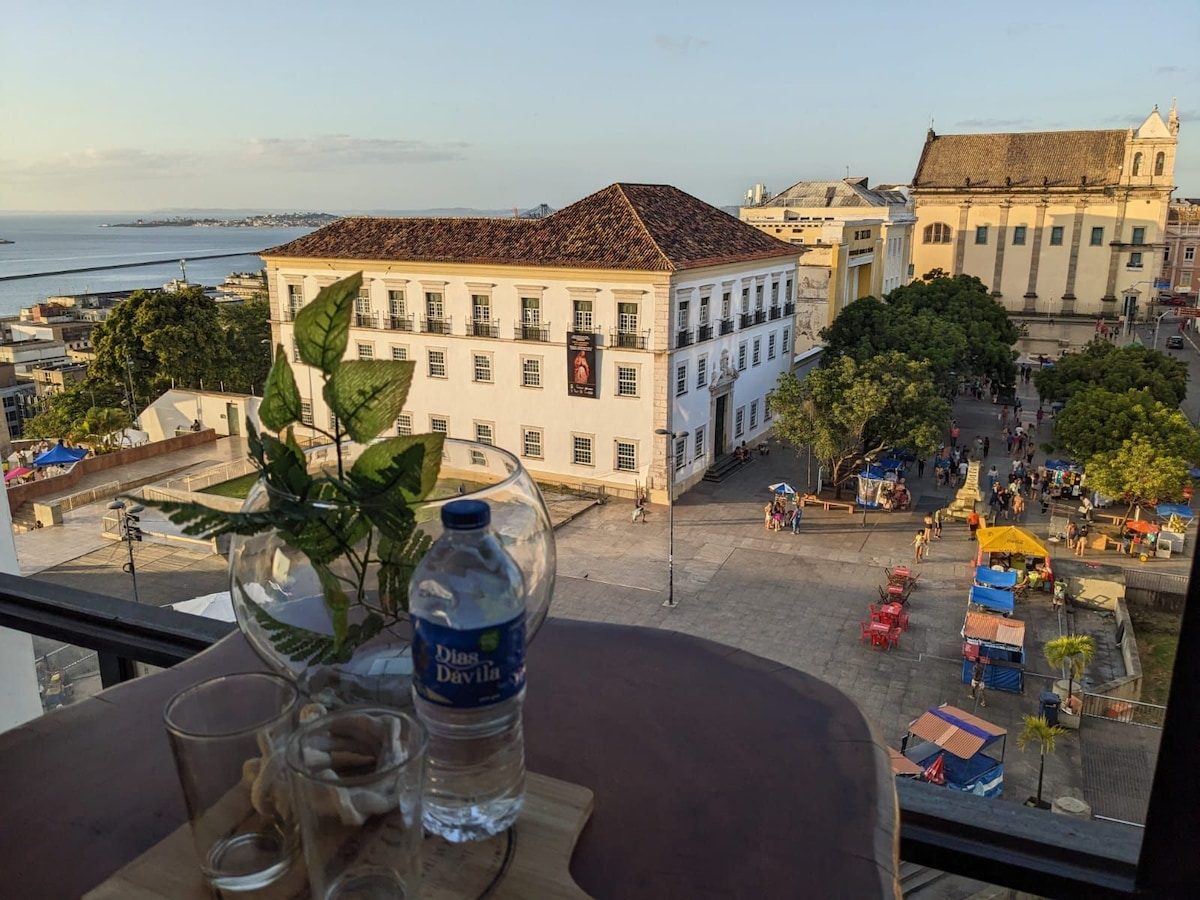 A glass of water and plants on a table by the window, looking down on a square of colonial buildings and the bay