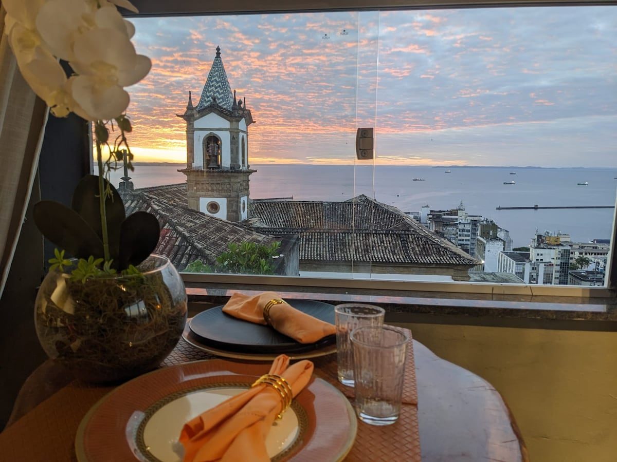 A table laid for two at sunset by the window, a church bell tower and the Bay of All Saints beyond