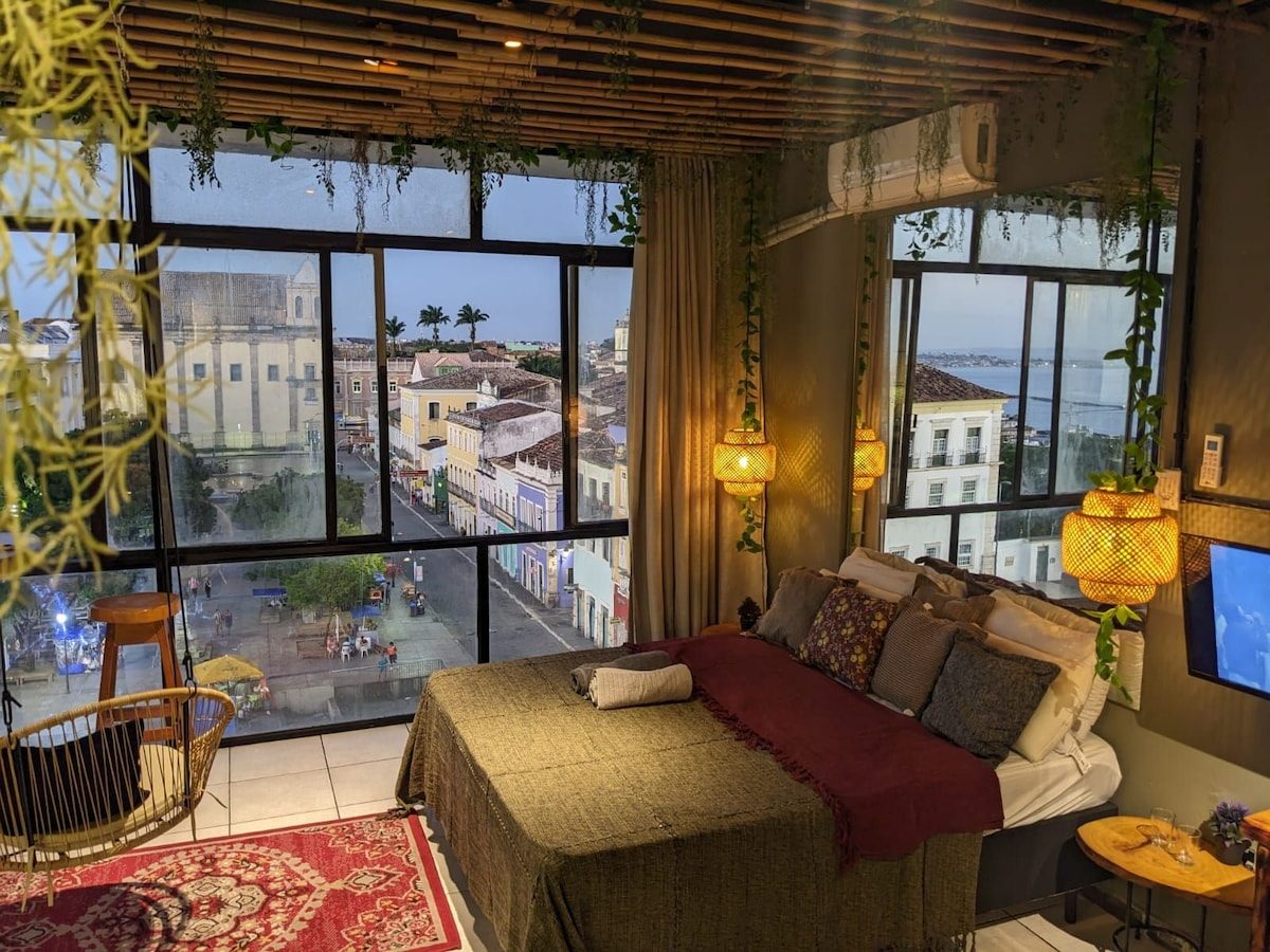 Bed with cushions and a red throw beneath a slatted ceiling, wide windows on the old town