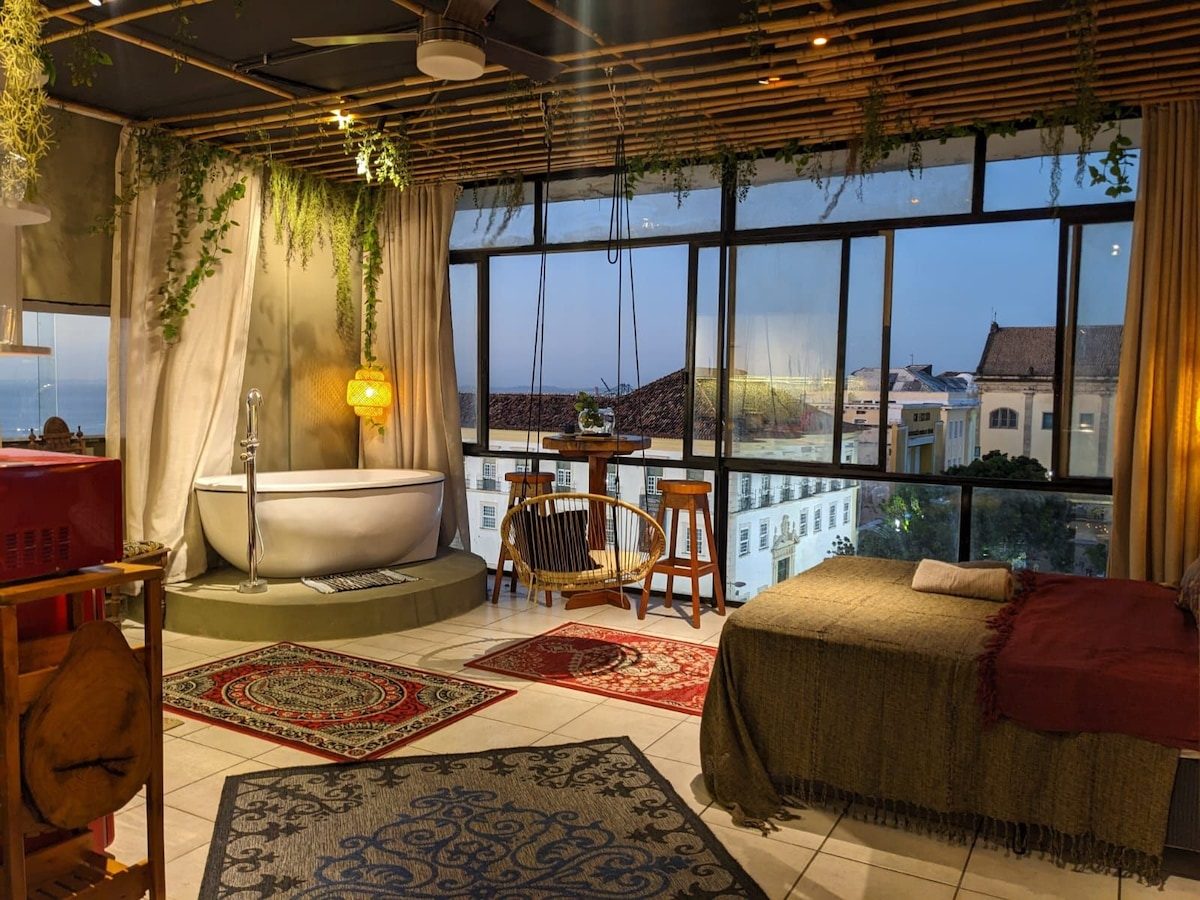 Suite at dusk with a freestanding tub on a platform by tall windows, a bed with a red throw and patterned rugs, the old town lit outside