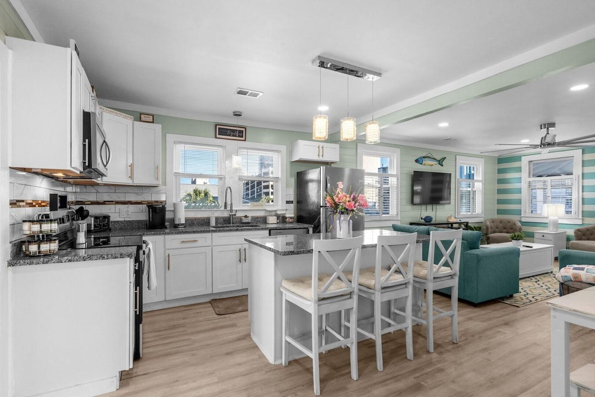 Bright white kitchen at Wait and Sea with a granite island, breakfast-bar seating and pendant lighting, open to the living room