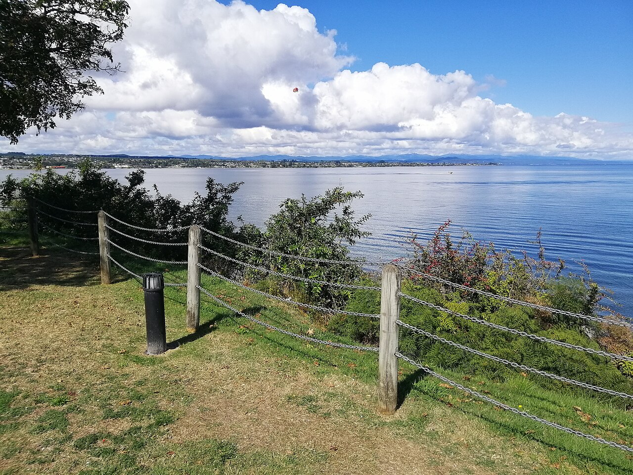 A grassy bank with a post-and-wire fence above a wide blue lake, with white clouds piled along the far shore.