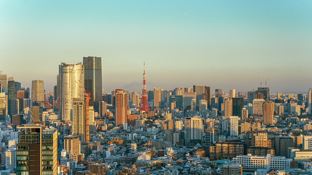 A dense cluster of skyscrapers in warm evening light, with a red and white lattice tower standing among them under a pale blue sky.