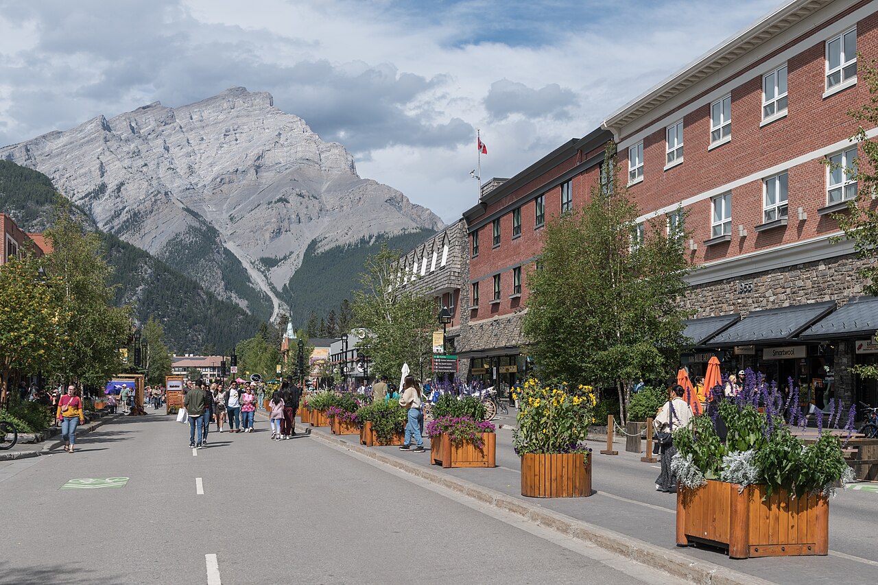 A wide street lined with buildings and trees, with people walking along sidewalks and a large mountain in the background under a cloudy sky.