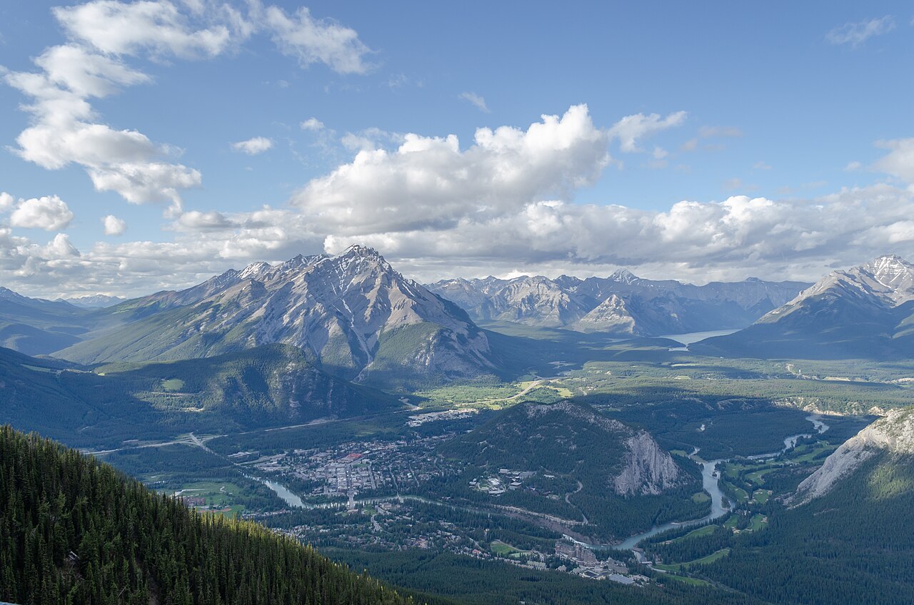 An aerial view of a small town nestled in a valley, surrounded by large, tree-covered mountains under a partly cloudy sky.