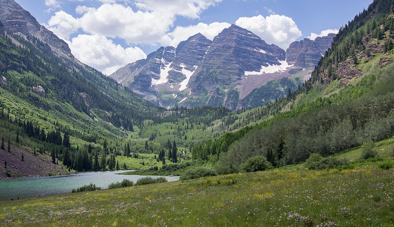 A wide shot of two prominent, snow-capped mountains rising above a green valley with a lake in the foreground.