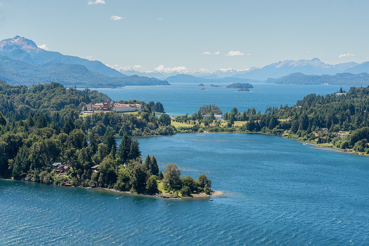 An aerial view of a large lake with several islands and peninsulas covered in green trees, surrounded by mountains under a blue sky.