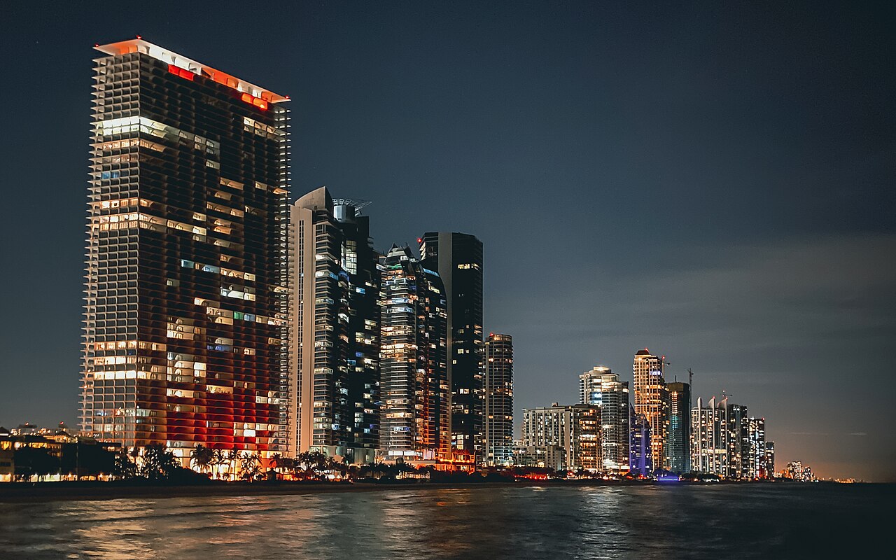 A nighttime view of a city skyline with numerous illuminated skyscrapers reflecting in the dark water.