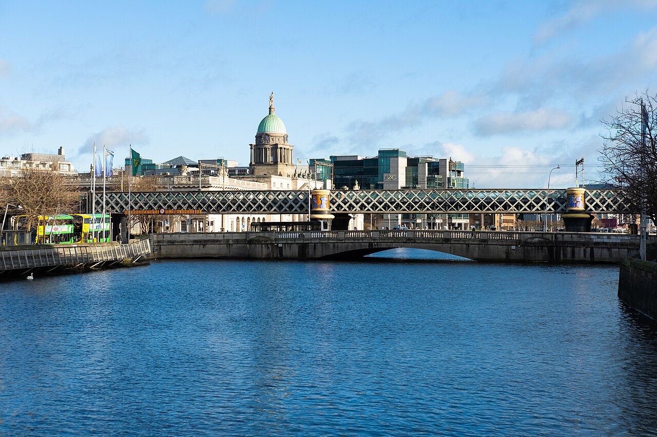 A river crossed by a low stone bridge and an iron railway bridge, with a domed building rising above the rooftops behind.