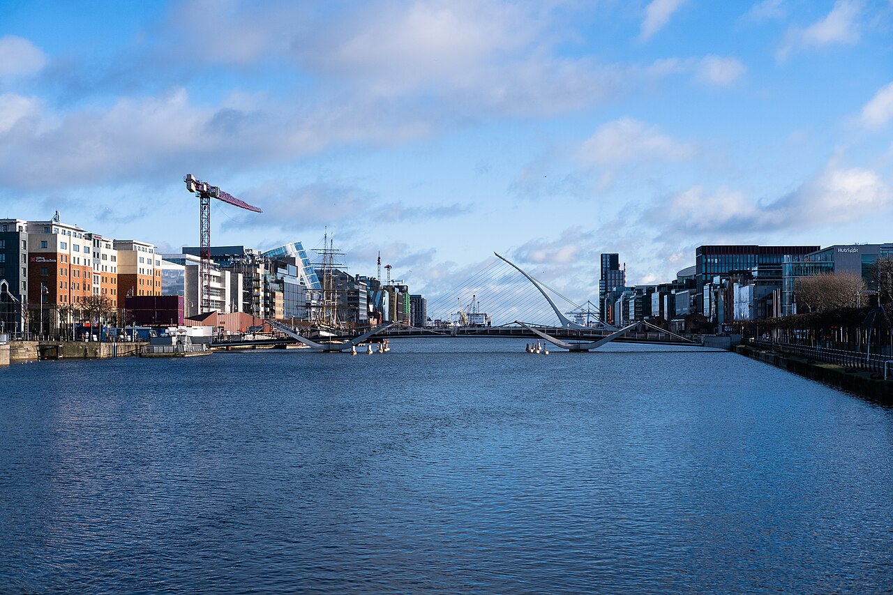 A broad, calm river between modern quayside buildings, with a white cable-stayed bridge and a crane in the distance under a blue sky.
