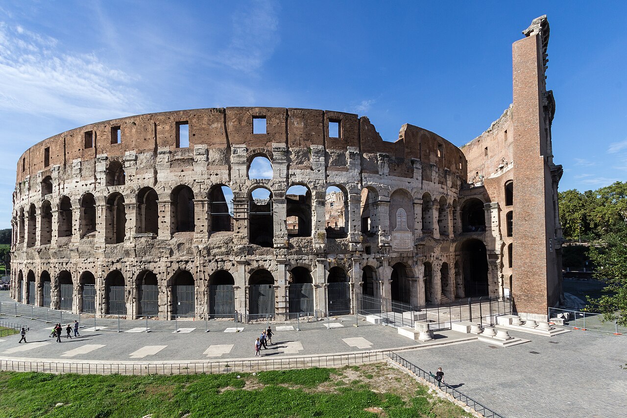 The curved outer wall of an ancient stone amphitheatre with tiers of arches, seen across a paved square with a few distant visitors.