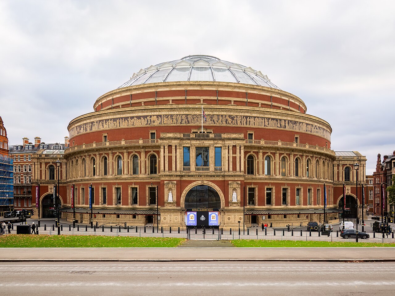 A large circular red-brick concert hall with a glass and iron dome and a decorative frieze around its upper wall, under an overcast sky.