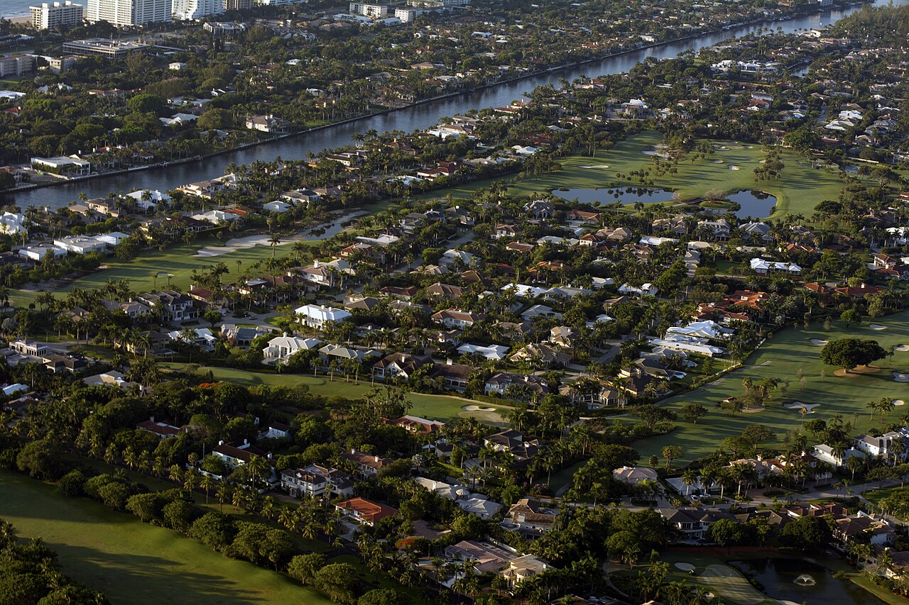 An aerial view of a residential area with many large houses, green golf courses, and a canal.