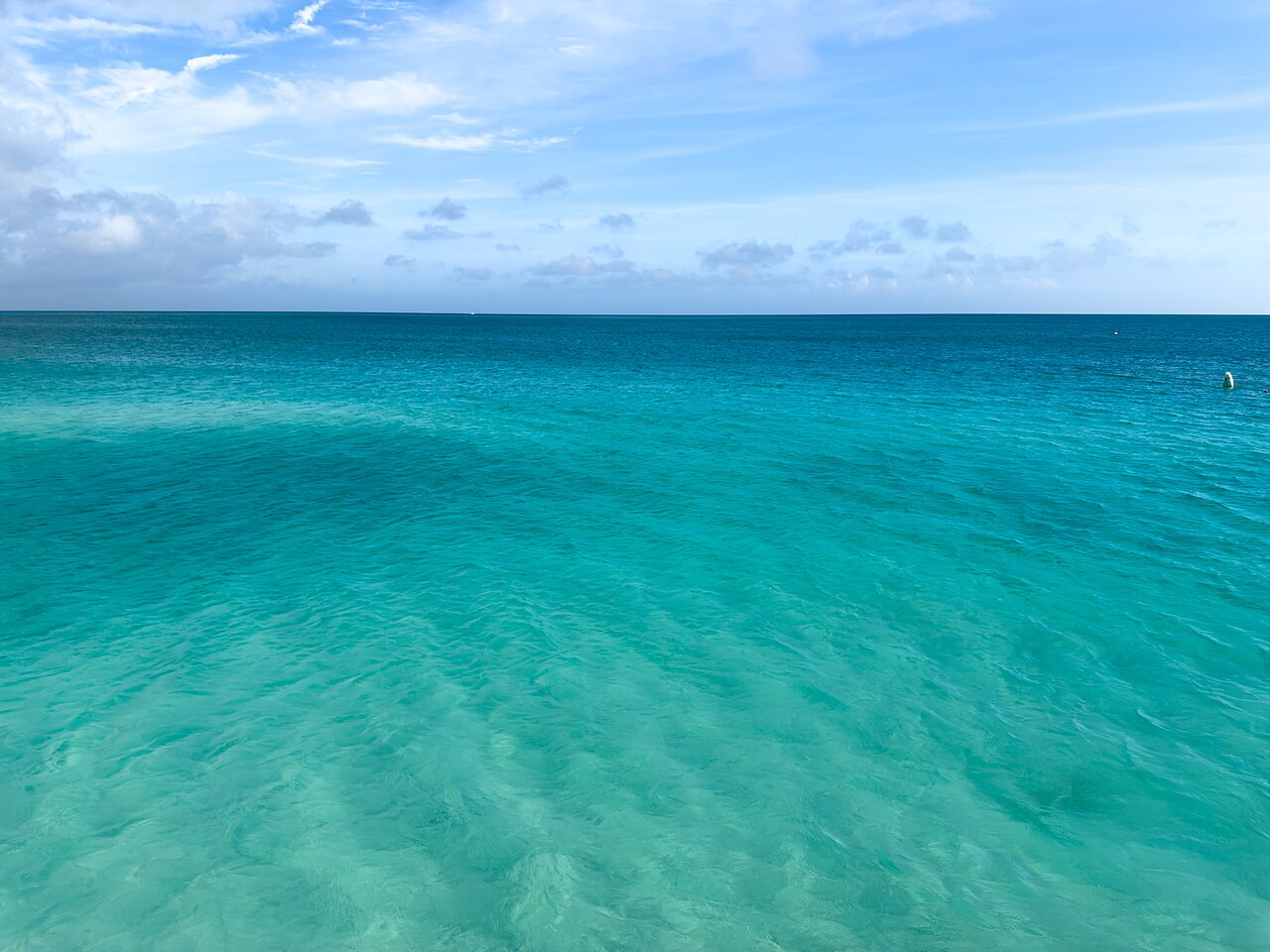 A wide shot of calm, clear turquoise ocean water with subtle ripples, extending to a light blue horizon under a partly cloudy sky.