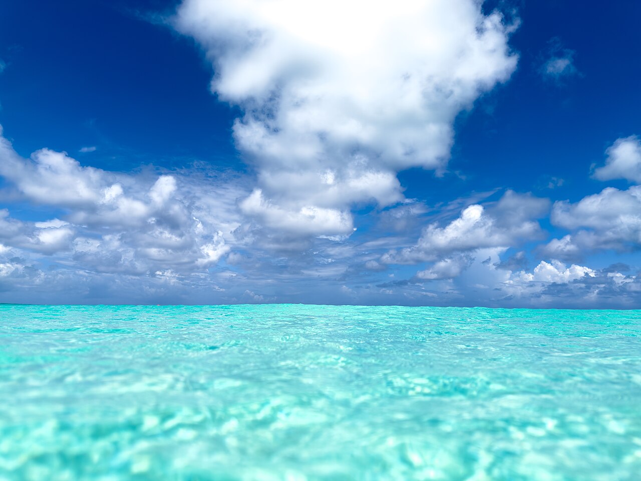 A low-angle view of incredibly clear, shallow turquoise water extending to the horizon, with large white cumulus clouds against a deep blue sky above.