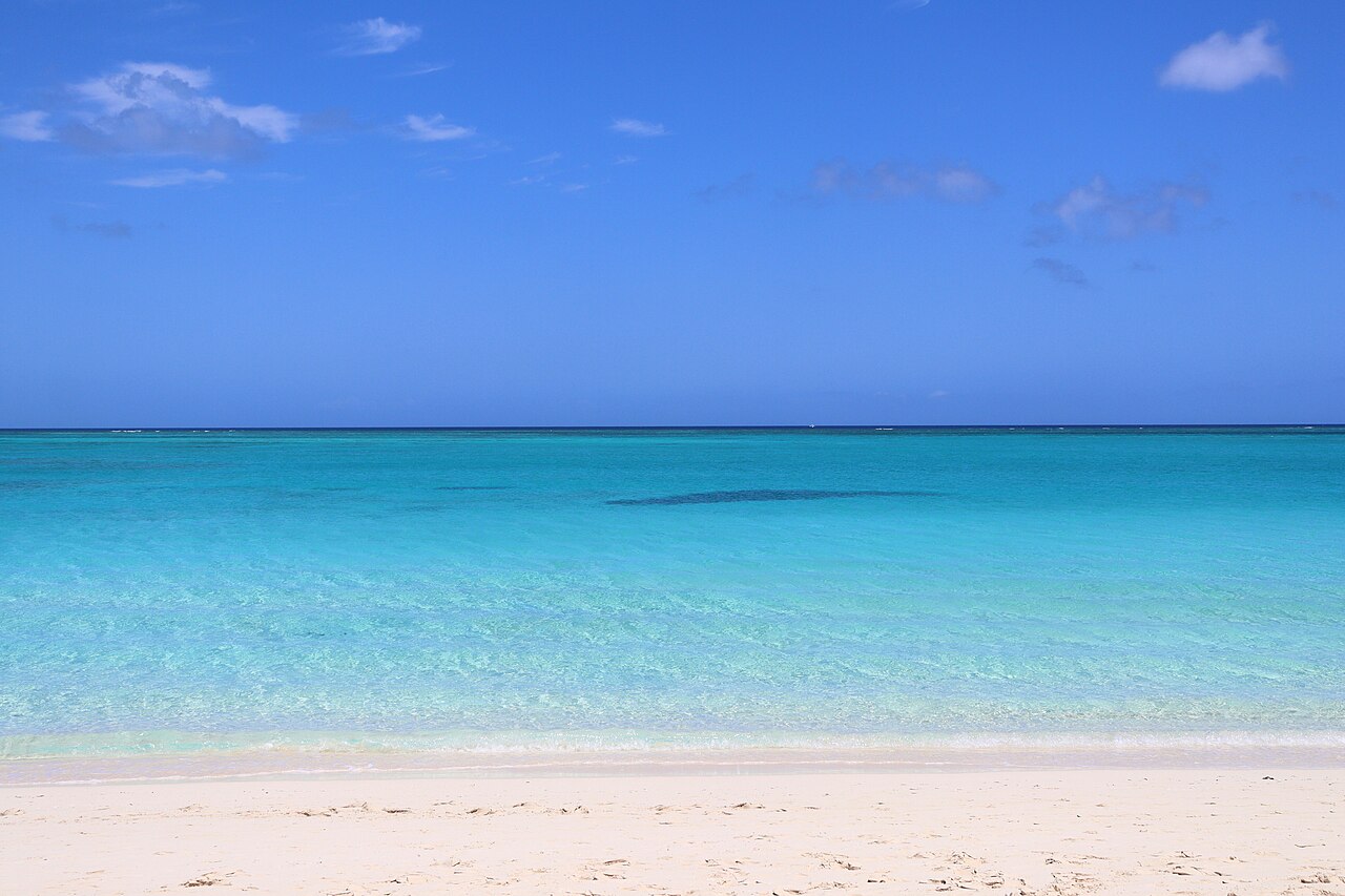 A wide shot of a pristine white sand beach meeting clear turquoise water under a bright blue sky with a few wispy clouds.