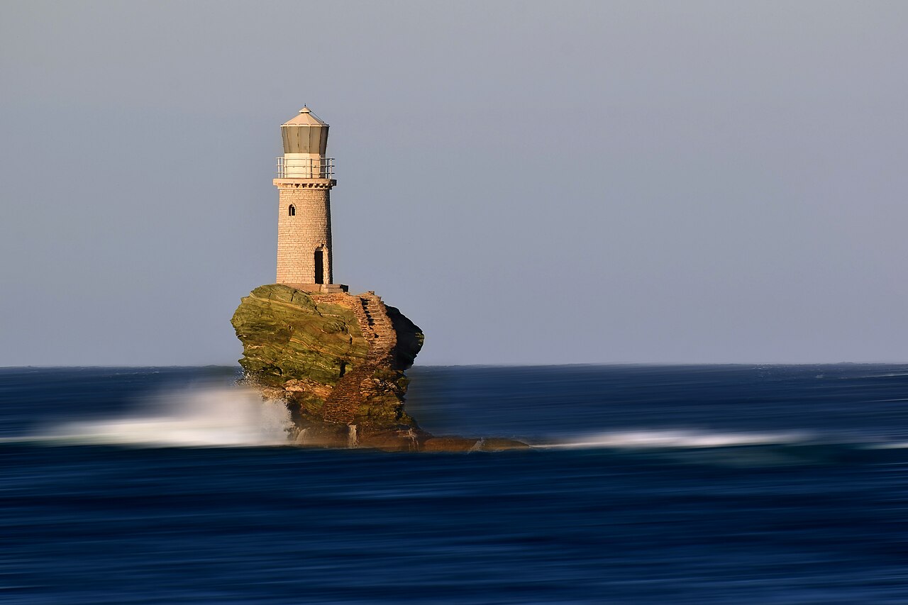 A slim stone lighthouse standing on a solitary weathered rock in the sea, the water smoothed to a blur by a long exposure.