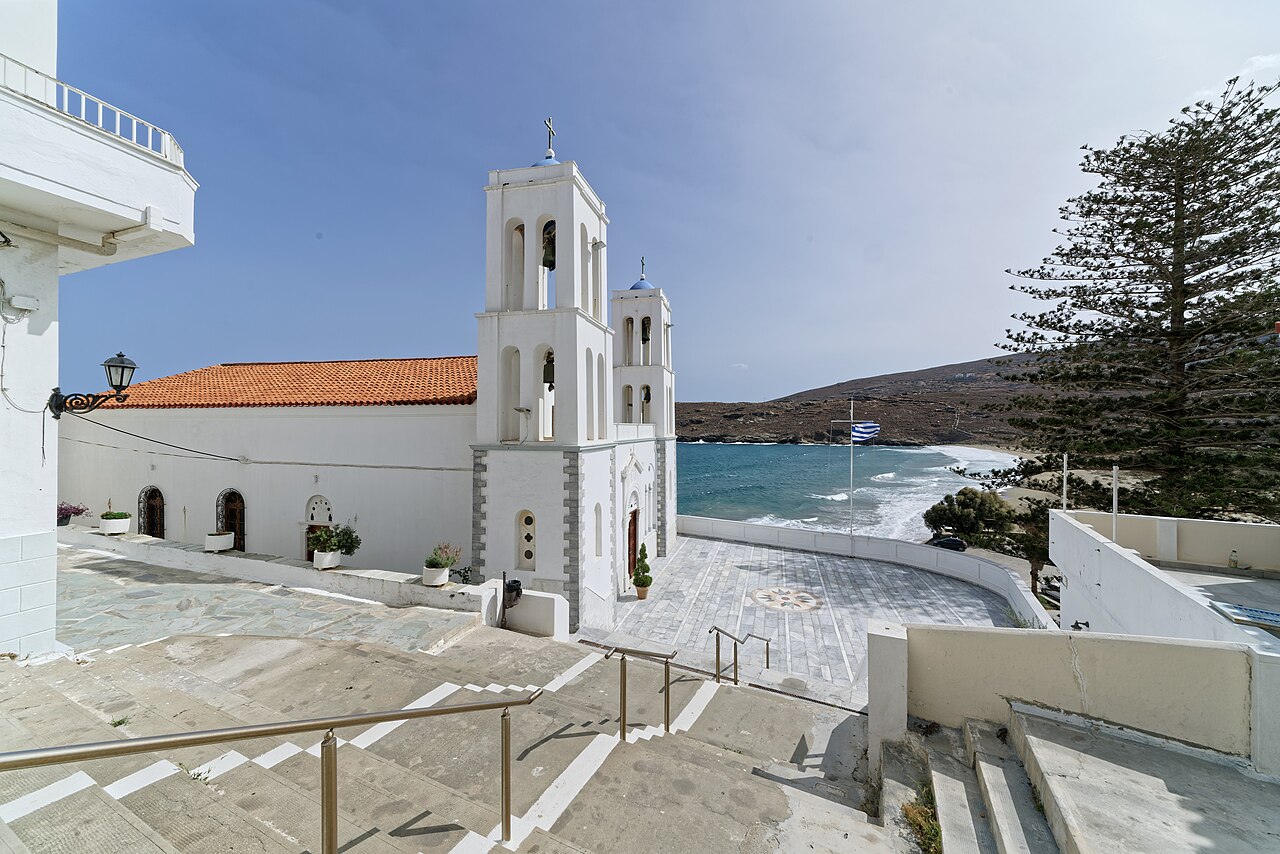 A whitewashed church with a tiered bell tower and a red tiled roof beside stone steps, with the sea and hills beyond.